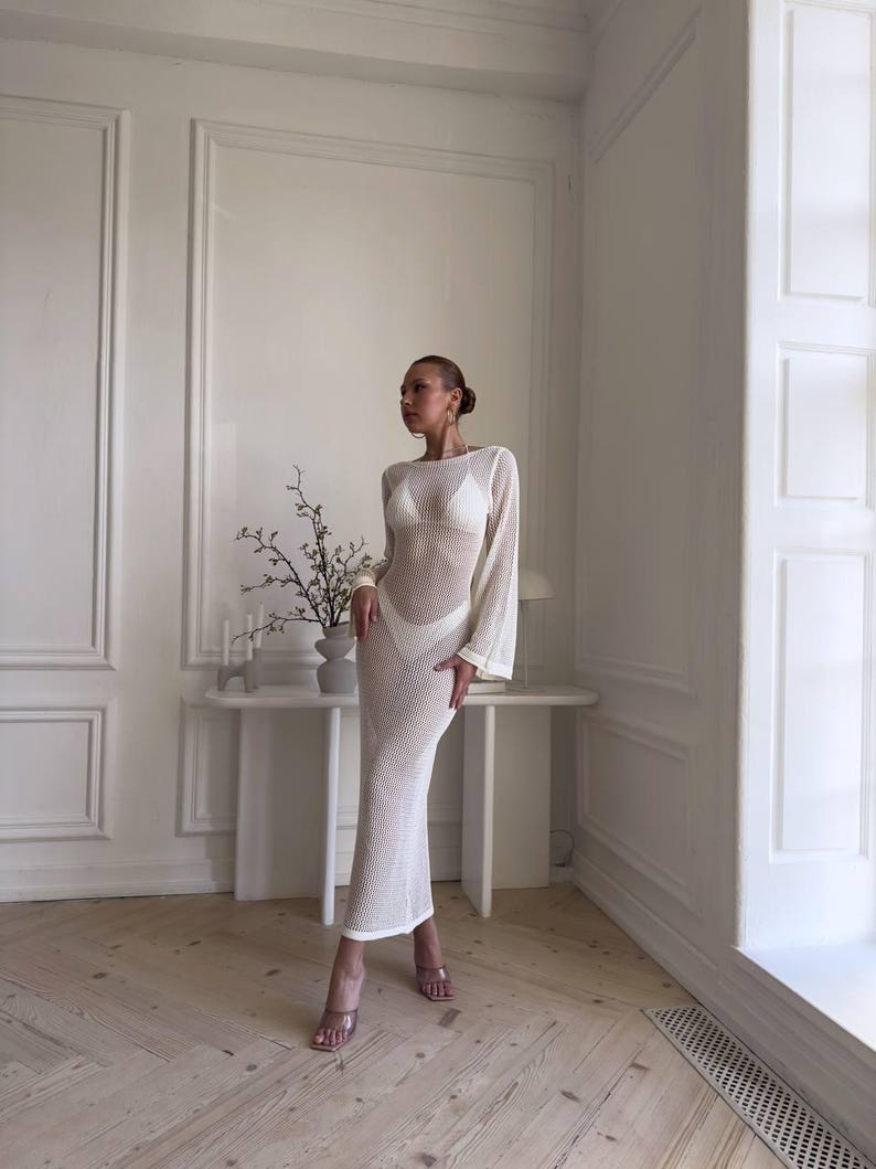 May include: A white, long-sleeved, open-weave maxi dress. The dress has a fitted silhouette and a sheer appearance. The model is wearing a white bikini underneath. The dress has a round neckline and flared sleeves. The model is wearing clear heels.