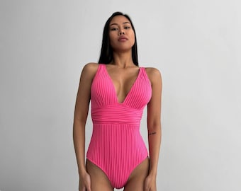 Peut inclure: Un maillot de bain rose côtelé avec un décolleté en V profond et des bretelles fines. Le maillot de bain a une jambe haute et un dos bas.