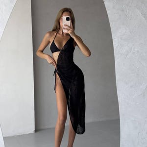 Op de afbeelding: Een zwarte, doorschijnende maxi-jurk met een zijsplit en een strikdetail in de taille. De jurk is gemaakt van een lichtgewicht, ademende stof en is perfect voor een dagje op het strand of een avondje uit.