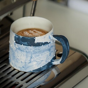 Pode incluir: Uma caneca de cerâmica com um design de retalhos azul e branco, cheia de café e arte latte. A caneca tem uma alça azul escura e está em uma máquina de café expresso de aço inoxidável.
