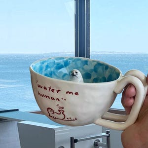May include: A quirky ceramic mug featuring a seal figurine. The mug has a white exterior with blue detailing and the red text "water me human". The mug is held in a hand, with a scenic ocean view in the background.
