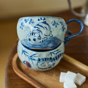 May include: Two stacked ceramic mugs with blue and white cat illustrations. The top mug has a handle and a cat design on the side. The bottom mug has a similar cat design. The mugs are on a wooden tray with sugar cubes.
