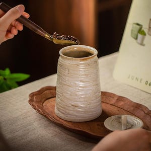 May include: A beige ceramic jar with a textured surface, sitting on a wooden tray. A small scoop with a wooden handle and a brass scoop is filled with coffee beans and is held above the jar. A small lid sits next to the jar.