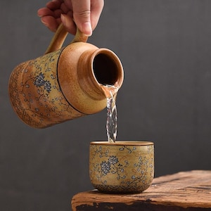 May include: A ceramic jug with a handle pours water into a matching cup. Both the jug and cup feature a beige background with blue floral designs and brown accents. The water stream is clear.