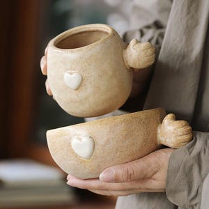 May include: Two beige ceramic planters, each with a white heart and a hand-shaped handle. The planters have a speckled texture and a rounded shape. One planter is held above the other.
