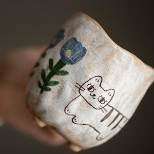 Könnte beinhalten: Handgefertigte Keramiktasse mit einer skurrilen Katzenillustration und blauen floralen Akzenten. Die Tasse hat eine strukturierte, cremefarbene Oberfläche und eine einzigartige, unregelmäßige Form. Die Katze ist in einem einfachen Cartoon-Stil gezeichnet.