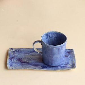 May include: A blue ceramic mug with a handle sits on a matching rectangular tray. The mug and tray have a textured, glazed finish with variations in the blue color. The set is designed for serving beverages.