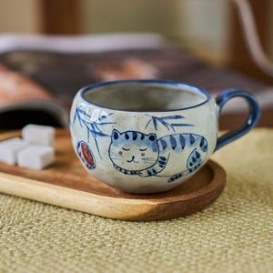 Könnte beinhalten: Eine Keramik-Tasse mit blauem Henkel und handgemaltem Katzenmotiv. Die Tasse ist weiß mit blauen Akzenten, und die Katze ist schlafend dargestellt. Die Tasse steht auf einem Holztablett mit Zuckerwürfeln.