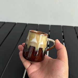 May include: A small ceramic mug with a unique glaze. The mug features a gradient of colors, including brown, tan, and white, with a dripped glaze effect. It has a small handle and a rounded shape, perfect for coffee or tea.