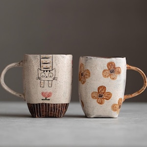 Puede incluir: Una taza de cerámica blanca con un asa negra y el texto "But First, Coffee" en negro y negrita. La taza está sobre una superficie blanca.