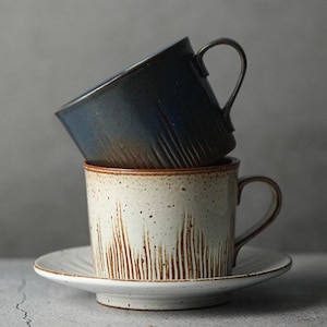 May include: Two ceramic mugs stacked on a white saucer. The top mug is dark blue with a textured surface, and the bottom mug is off-white with brown vertical lines. The saucer is also white with a slightly raised edge.