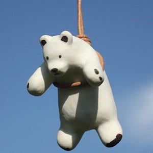May include: A white ceramic polar bear figurine with black accents on its paws, ears, and nose, suspended by a brown rope against a clear blue sky. The bear is in a playful pose, with its arms outstretched.