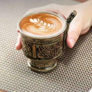 May include: A ceramic coffee cup with a decorative handle and base, filled with a latte featuring a leaf design in the foam. The cup has an aged, bronze-like finish with embossed patterns. The cup is held by a hand.