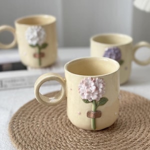 May include: Cream ceramic mugs with a sculpted hydrangea flower design. The mugs have a round handle and a detailed floral accent in pink, green, and brown. The mugs are set on a woven mat.