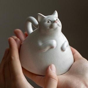 May include: A white ceramic cat-shaped mug with a rounded body, small paws, and a curved handle. The cat's face has detailed features, including a pink nose and expressive eyes. The mug is held in two hands.