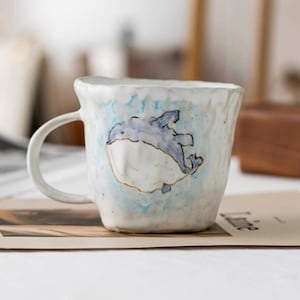Puede incluir: Una taza de cerámica hecha a mano con un diseño caprichoso de ballena. La taza tiene una base blanca con un fondo de acuarela azul y blanco. La ballena está representada en tonos azules. La taza tiene un asa curva.