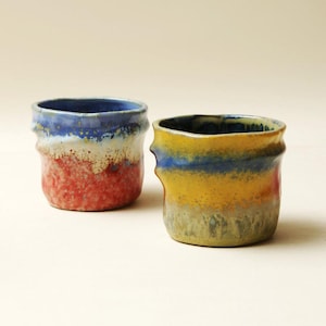 May include: Two ceramic cups with irregular, wavy rims and textured surfaces. The left cup features a blue top, a white middle band, and a red base. The right cup displays a blue top, a yellow middle section, and a muted green base with glaze.