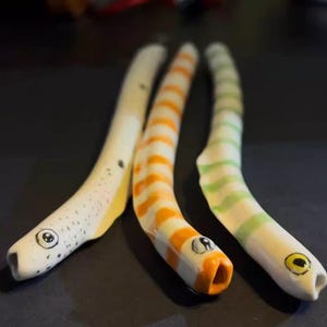 Puede incluir: Tres esculturas de cerámica de anguilas caprichosas. Una es blanca con manchas negras y detalles amarillos, otra es de rayas naranjas y blancas, y la tercera es blanca con rayas verdes. Cada una tiene una cara de dibujos animados.