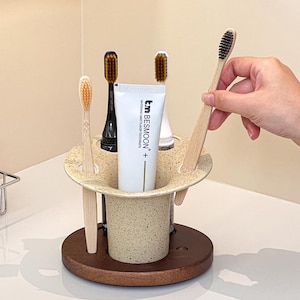May include: A beige speckled toothbrush holder with a dark brown circular wooden base. The holder contains a white tube of Besmoon whitening mint toothpaste and four toothbrushes with varying handle colours and bristle types.
