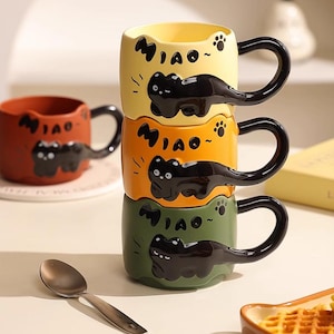 May include: A stack of three ceramic mugs in yellow, orange, and green, each with a black cat design and the word "Miao." The mugs have black handles and paw print details. A red mug with the same design is also visible.