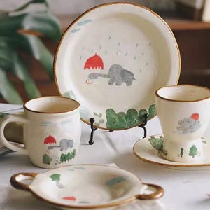 May include: A cream-colored ceramic dish set featuring an elephant design. The set includes a large plate, a mug, a small bowl, and a cup. Each piece has a brown rim and a whimsical illustration of an elephant holding a red umbrella in the rain.