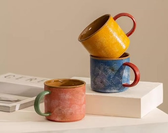 May include: Three ceramic mugs in a stack. The top mug is yellow with a red handle, the middle mug is blue with a red handle, and the bottom mug is red with a green handle. The mugs have a textured, speckled finish.