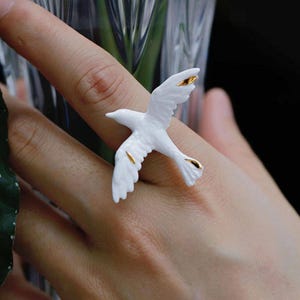 May include: A white bird ring with gold accents. The ring features a detailed design of a bird in flight, with gold highlights on the wings and tail. The ring is a unique piece of jewelry.