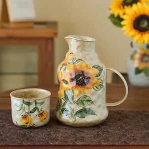 May include: A ceramic pitcher and cup set with a sunflower design. The pitcher has a handle and is painted with a large yellow sunflower. The cup features a smaller floral design. The set is on a brown surface.
