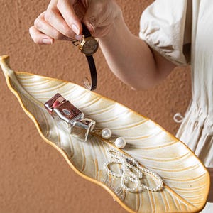 以下が含まれることがあります： クリームとゴールドの色合いの葉の形をした装飾トレイ。小さな時計、香水瓶、パールのジュエリーが置かれています。トレイはテクスチャ加工された表面で、小物を入れるように設計されています。