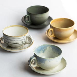 Puede incluir: Cuatro tazas y platillos de cerámica en varios colores: verde, amarillo, azul y blanco. Las tazas tienen formas redondeadas con asas y se asientan sobre platillos a juego. La taza blanca tiene líneas negras. Las tazas están sobre una superficie blanca.