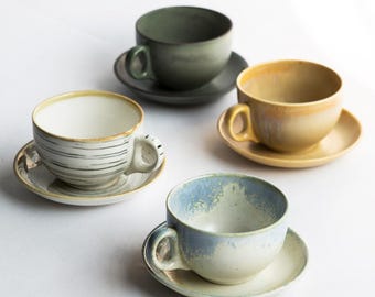 May include: Four ceramic teacups and saucers in various colors: green, yellow, blue, and white. The cups have rounded shapes with handles and sit on matching saucers. The white cup has black lines. The cups are on a white surface.