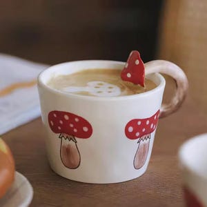 May include: A white ceramic mug with a mushroom design. The mug features red mushrooms with white polka dots and a brown stem. The handle is designed to look like a mushroom stem. The mug is filled with a creamy beverage and a mushroom-shaped design on top.