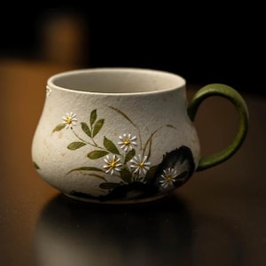 May include: A cream-coloured ceramic mug with a green handle. The mug features a floral design of white daisies and green leaves. The mug has a rounded shape and a textured surface. The handle is a dark green colour.