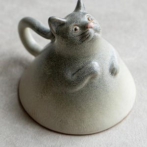 以下が含まれることがあります： 斑点のある質感の丸い灰色の猫の形をした陶器のマグカップ。猫にはピンクの鼻、大きな目、胸元に上げられた小さな前足があります。後ろからは尻尾のような湾曲した持ち手が伸びています。