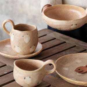 May include: A collection of handmade ceramic tableware in a warm beige color. The set includes a mug with a handle, a saucer, a bowl with a handle, and a plate. Each piece is decorated with white, cloud-like shapes and small black dots.