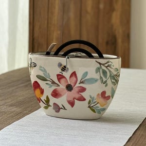 May include: A cream-colored ceramic incense holder with a wide, oval shape. The exterior features hand-painted watercolor illustrations of red and yellow flowers, green leaves, and a small bee. A black mosquito coil rests inside the holder.