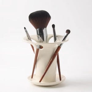 Può includere: Un supporto in ceramica bianca con parte superiore svasata e base integrata, caratterizzato da molteplici aperture circolari lungo il bordo. Quattro pennelli da trucco con manici in legno marrone scuro e ghiere argentate sono inseriti.