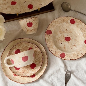 Puede incluir: Un juego de vajilla de cerámica con base color crema, textura marrón moteada y un patrón de manzanas rojas. El conjunto incluye una fuente grande, un cuenco, una taza y dos platos, todos con bordes irregulares y ondulados.