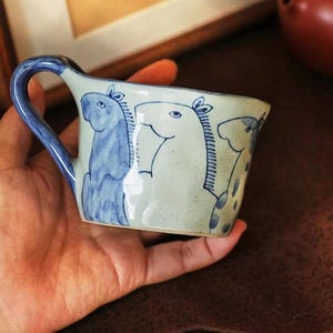 Puede incluir: Una taza de cerámica con un asa azul y un cuerpo blanco. La taza está decorada con ilustraciones azules de caballos. La taza está sostenida en la mano de una persona.