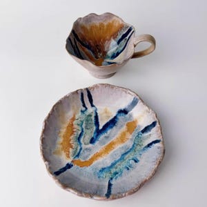 Könnte beinhalten: Ein Keramik-Tassen- und Teller-Set mit einer einzigartigen, organischen Form. Die Tasse hat einen Henkel und einen blütenblattartigen Rand. Teller und Tasse zeigen ein Design mit blauen, orangefarbenen und grünen Streifen auf hellem Hintergrund.