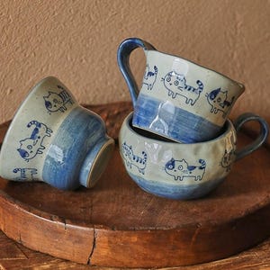 Puede incluir: Tres tazas de cerámica con ilustraciones de gatos azules y blancos. Las tazas están apiladas en una bandeja de madera. Las tazas tienen un borde y un asa azules. Las ilustraciones de gatos son de estilo caprichoso.