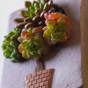 May include: A decorative succulent arrangement featuring green, orange, and brown plants. The plants are arranged in a pot with a brick-like design. The background is a light gray color, and the overall aesthetic is natural and earthy.