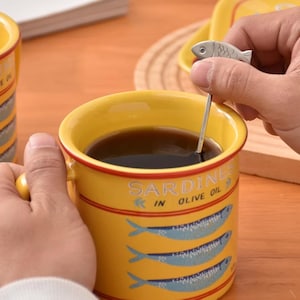 Puede incluir: Taza de cerámica amarilla con diseño de sardinas y el texto "Sardines in Olive Oil". Una pequeña cuchara con forma de pez revuelve el líquido oscuro en su interior. La taza tiene un asa amarilla y un borde rojo.