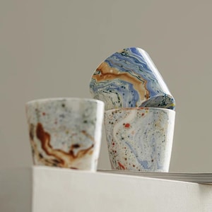 Può includere: Tre tazze in ceramica con un motivo marmorizzato con vortici di blu, marrone e bianco. Le tazze hanno una finitura maculata con piccoli punti rossi, neri e blu su tutta la superficie. Una tazza è posizionata di lato sopra un'altra tazza.