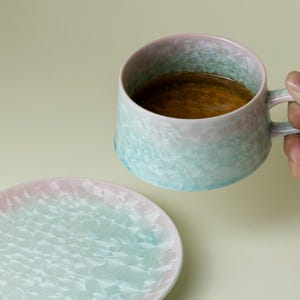 Könnte beinhalten: Ein Keramik-Tassen- und Untertassen-Set mit einem strukturierten, kristallinen Muster. Die Tasse ist mit einer braunen Flüssigkeit gefüllt. Die Farben gehen von Hellblau zu Rosa über. Die Untertasse ist rund und passt zum Design der Tasse.