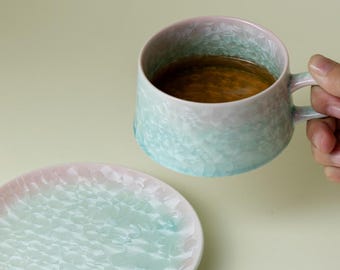 May include: A ceramic mug and saucer set with a textured, crystalline pattern. The mug is filled with a brown liquid. The colors transition from light blue to pink. The saucer is round and matches the mug's design.