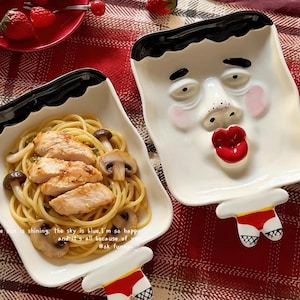 Puede incluir: Dos platos blancos de cerámica con forma de cara y borde negro, uno lleno de pasta y pollo, el otro vacío. Los platos tienen un diseño de cara de dibujos animados con labios rojos y rubor. Los platos tienen un asa con forma de ropa interior.