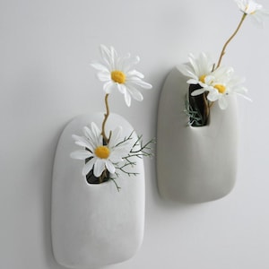May include: Two smooth, rounded, light grey wall-mounted vases with a matte finish. Each vase features a single opening at the top containing artificial white daisy flowers with yellow centres and green stems, mounted against a plain light grey wall.