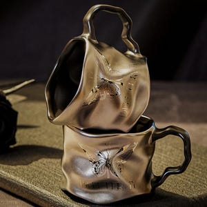 Könnte beinhalten: Zwei gestapelte, metallisch-goldene Kaffeetassen mit einer strukturierten, gewellten Oberfläche. Jede Tasse hat ein detailliertes Schmetterlingsdesign und das Wort "BUTTERFLY" auf der Seite eingraviert. Die Griffe haben eine einzigartige, wellenförmige Form.
