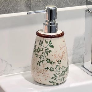 Könnte beinhalten: Ein Keramik-Seifenspender mit floralem Design. Der Spender hat eine cremefarbene Basis mit grünen und rosa Blumenmustern. Er hat einen braunen Rand und eine silberne Pumpe.